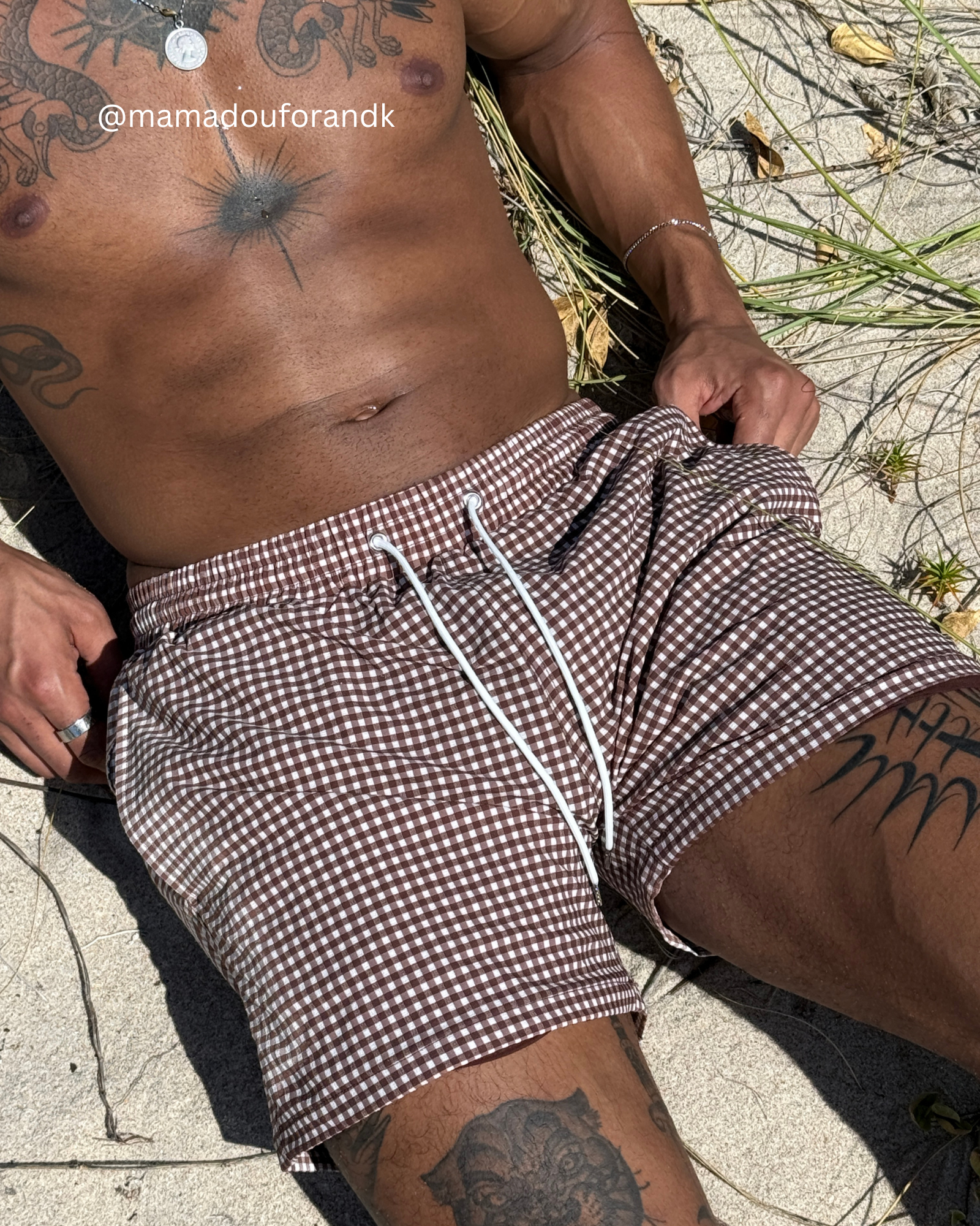 Person wearing checkered shorts sitting on a sandy surface with tattoos visible.