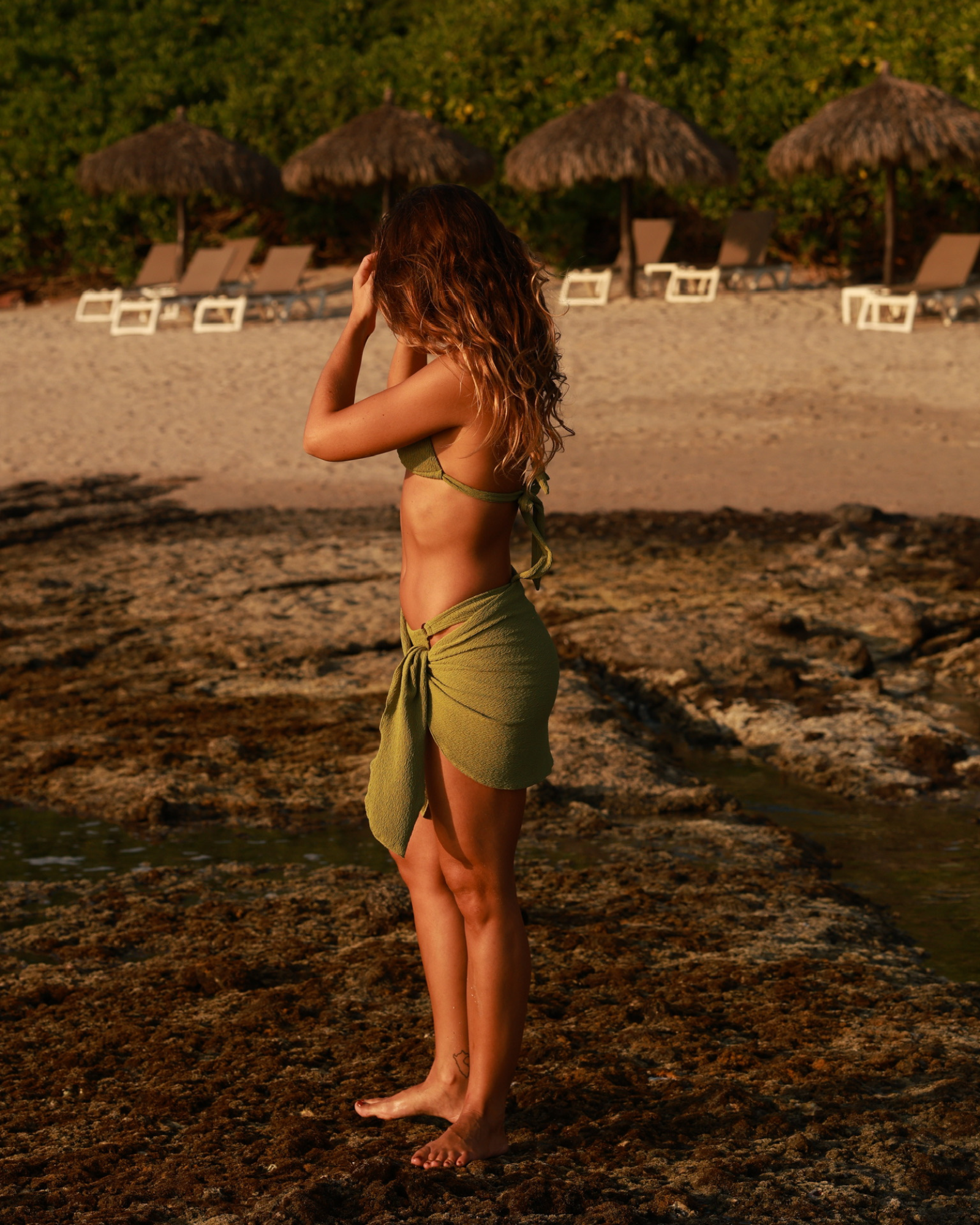 Woman in a green bikini and sarong standing on a beach with straw umbrellas in the background.
