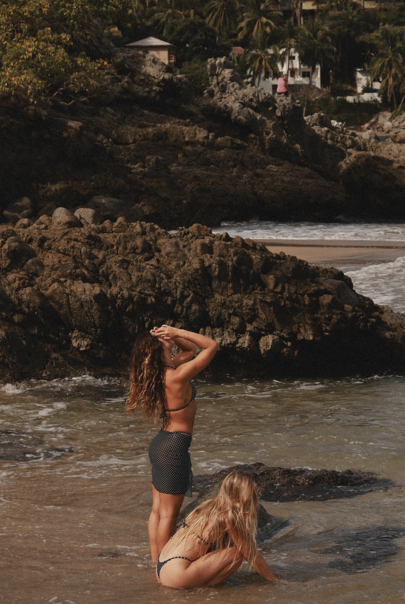 Two women at a beach with rocky cliffs and ocean waves, one wearing a short pareo.