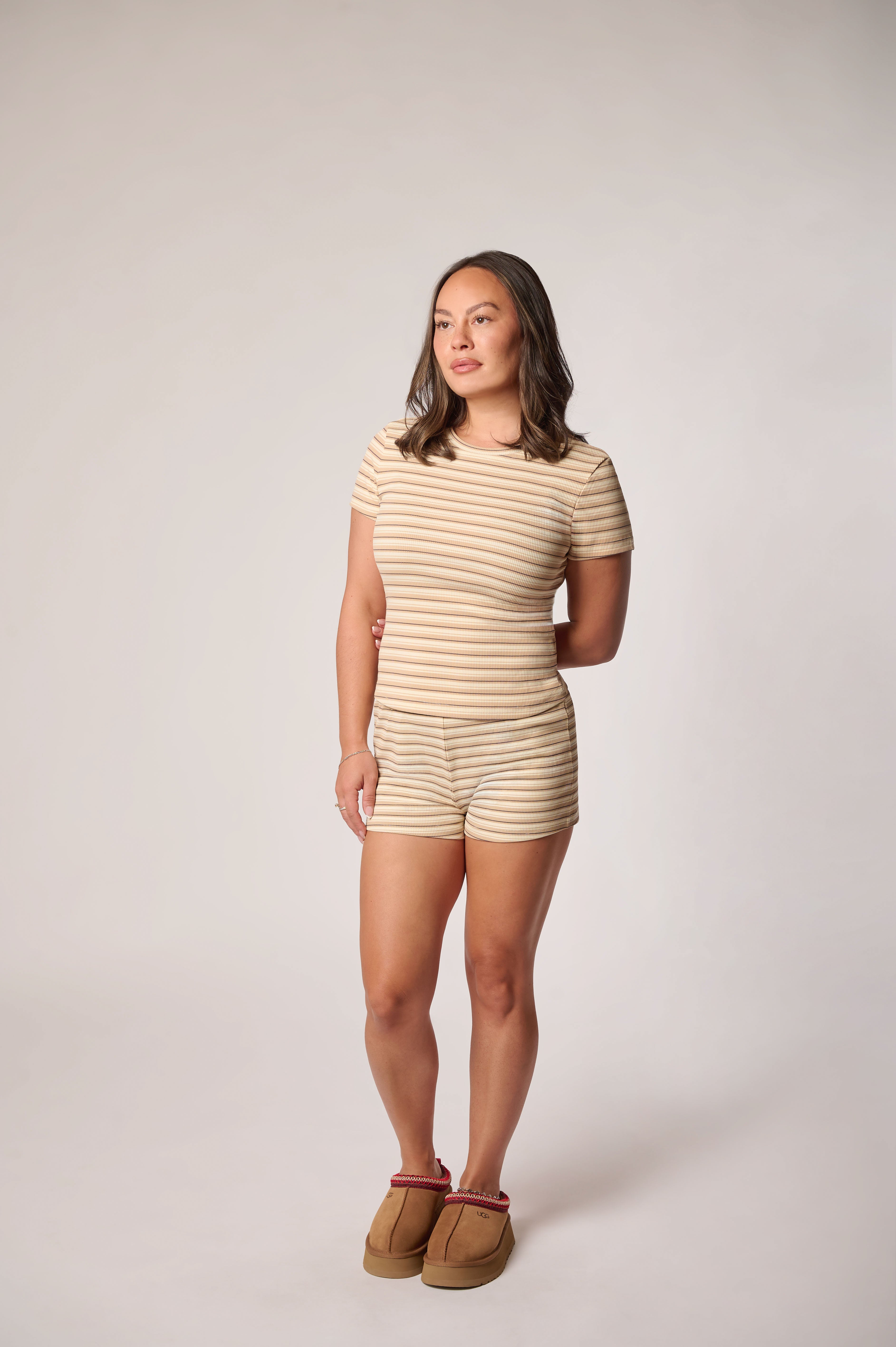 A woman modeling a beige and brown horizontally striped short-sleeve romper with matching shorts, paired with brown suede slippers