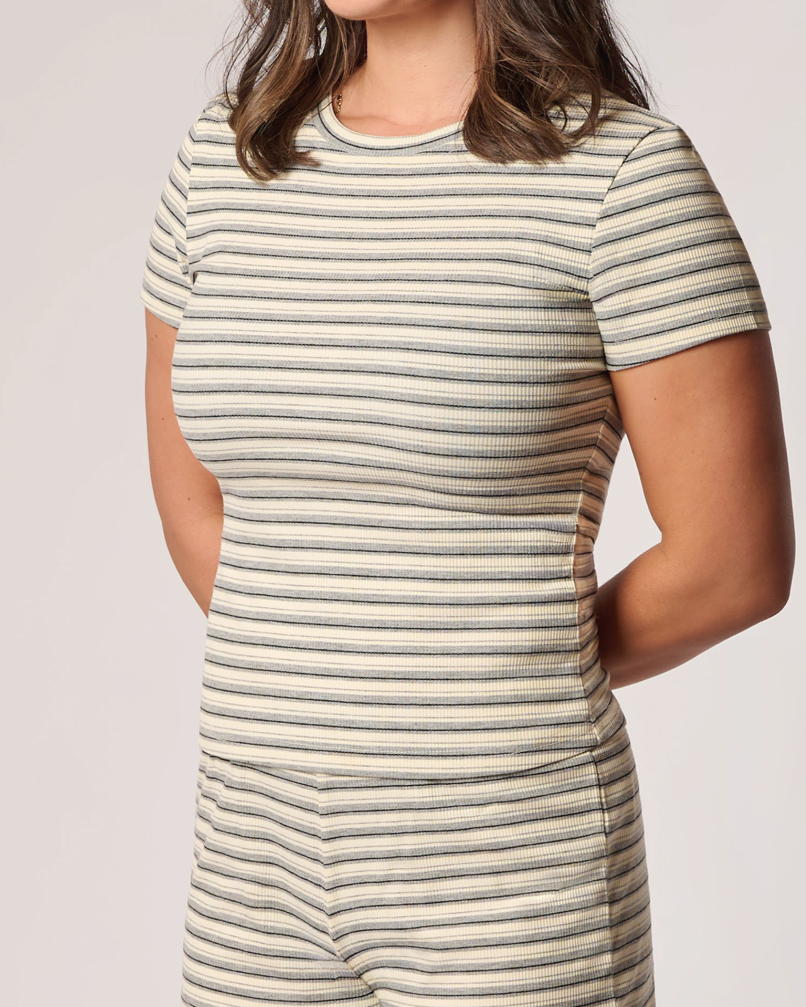 Close up of a woman wearing a short-sleeve ribbed knit top with thin horizontal stripes in beige, white, and dark gray, shown from the chest down against a plain light gray studio background.