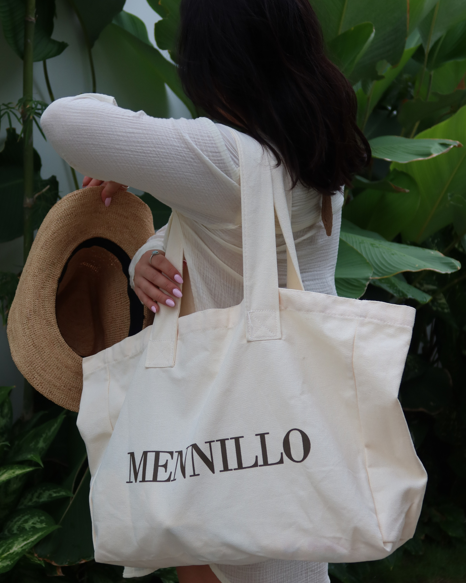 Women holding a beige tote bag with MENNILLO branding against a green leafy background