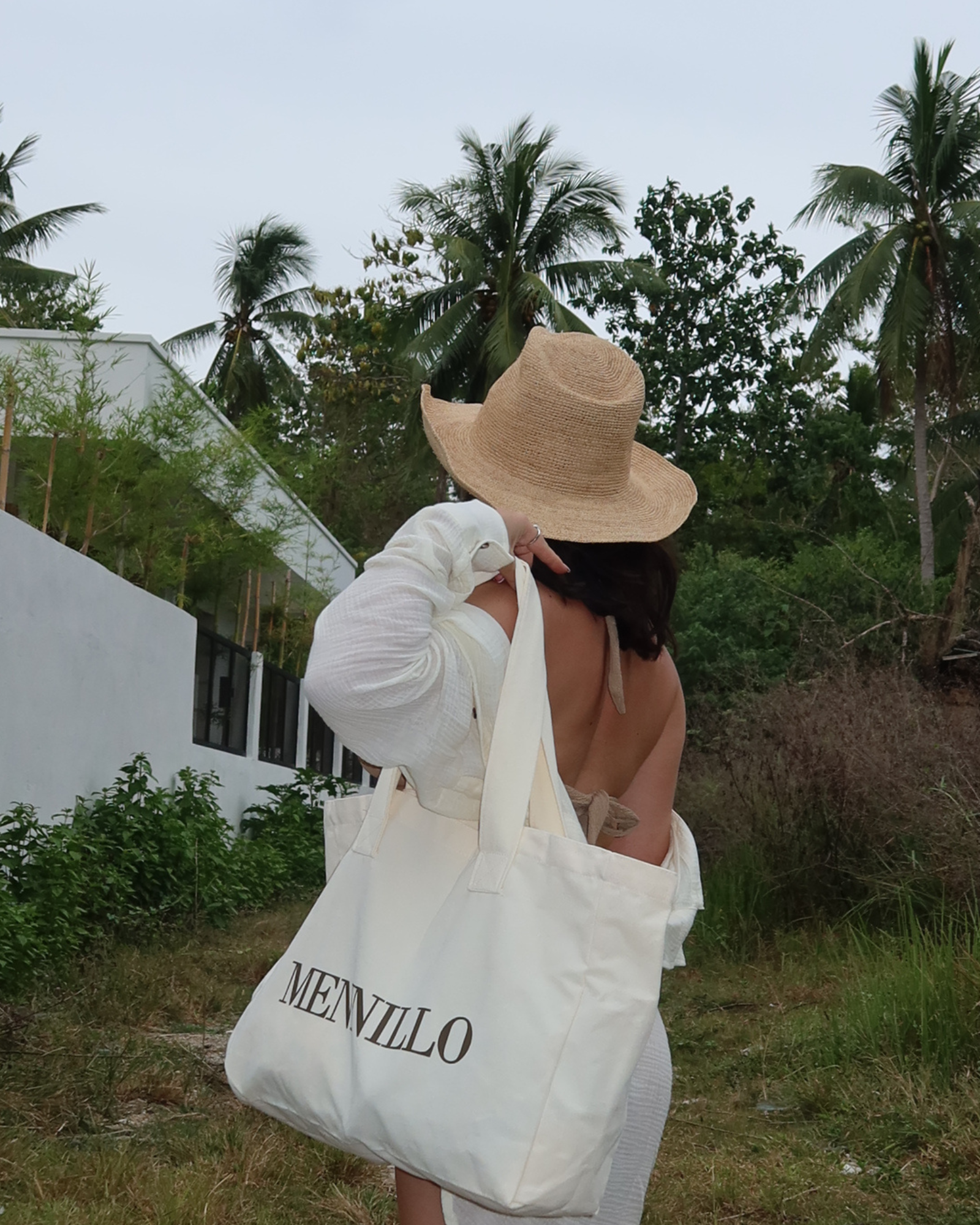 Person holding a white tote bag with MENNILLO branding in a tropical setting.
