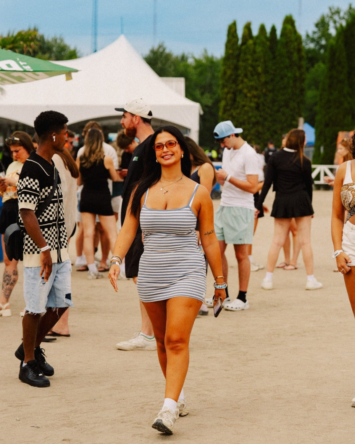 A woman wearing a gray and white striped mini dress walks through an outdoor crowd at a beachclub event.