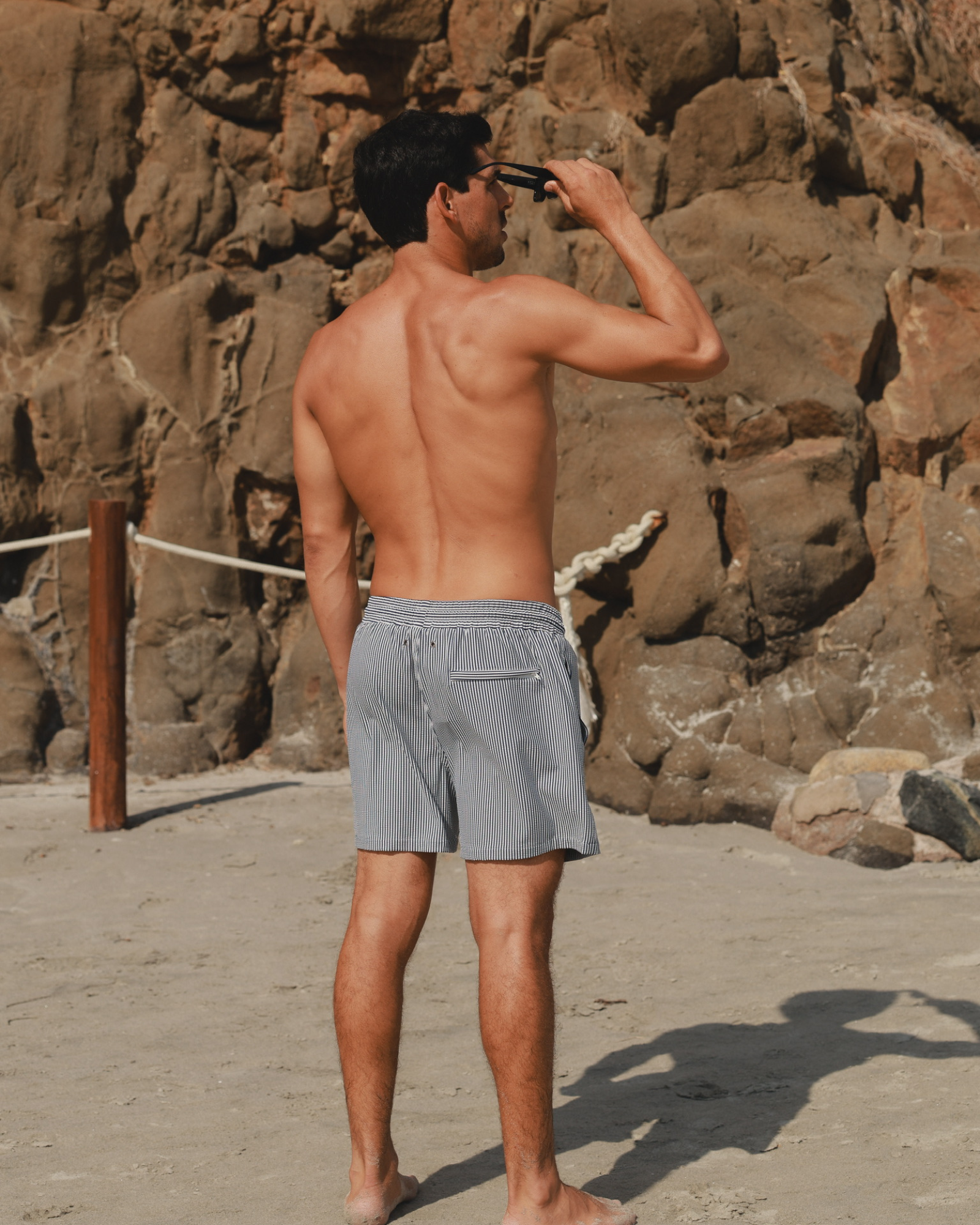 Man wearing striped swim shorts standing on a beach with rocky cliffs behind him.