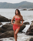 Woman in a red bikini skirt standing on a rocky beach with ocean and mountains in the background