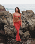 Woman in a red polka dot bikini  midi skirt standing on rocky terrain by the ocean.
