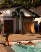 Two women in pink swimsuits by a pool with a house and palm tree in the background