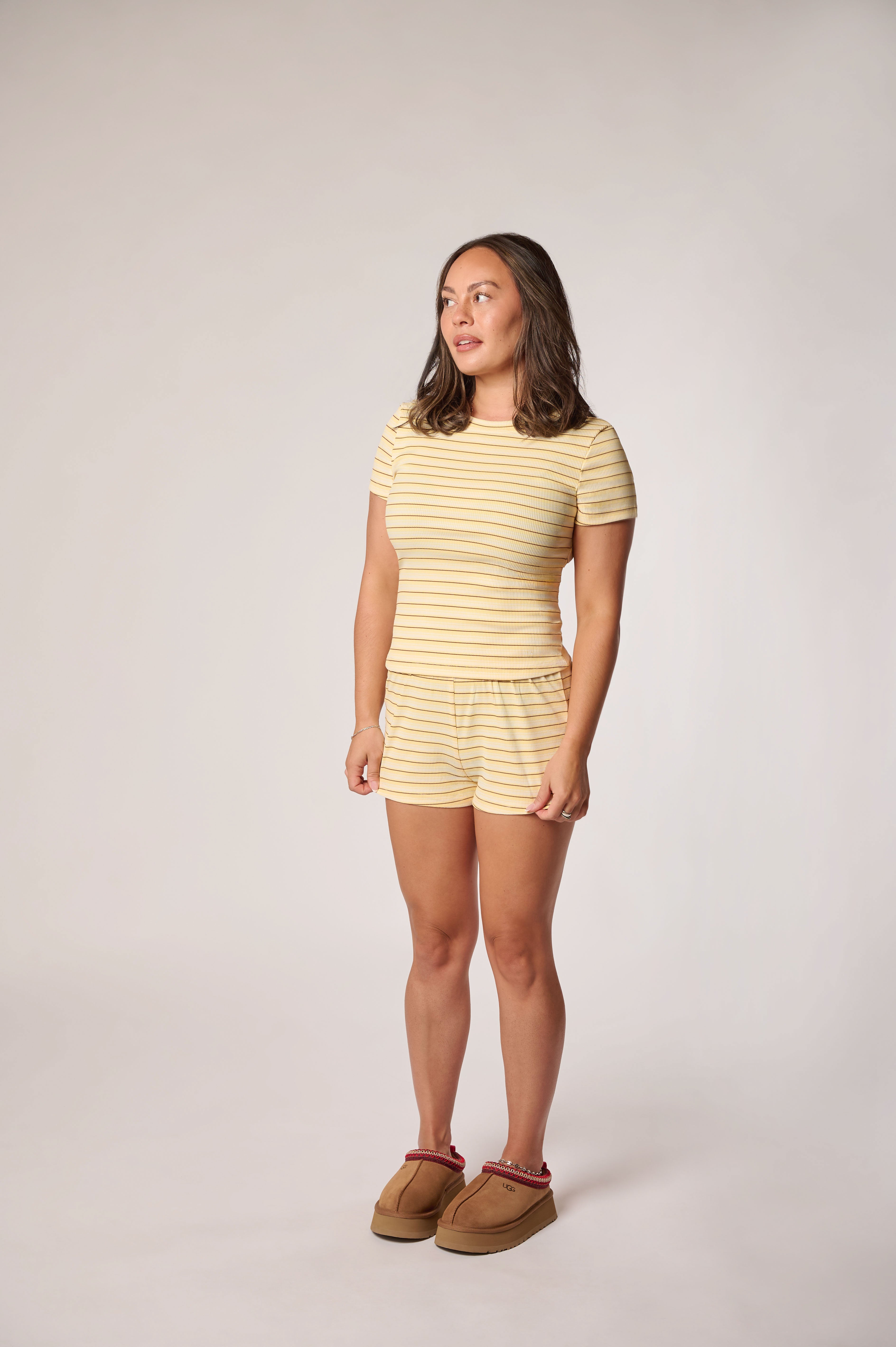 A woman modeling a matching two-piece loungewear set both featuring a horizontal yellow and white stripe pattern. She is also wearing brown suede slippers against a neutral light gray studio background.