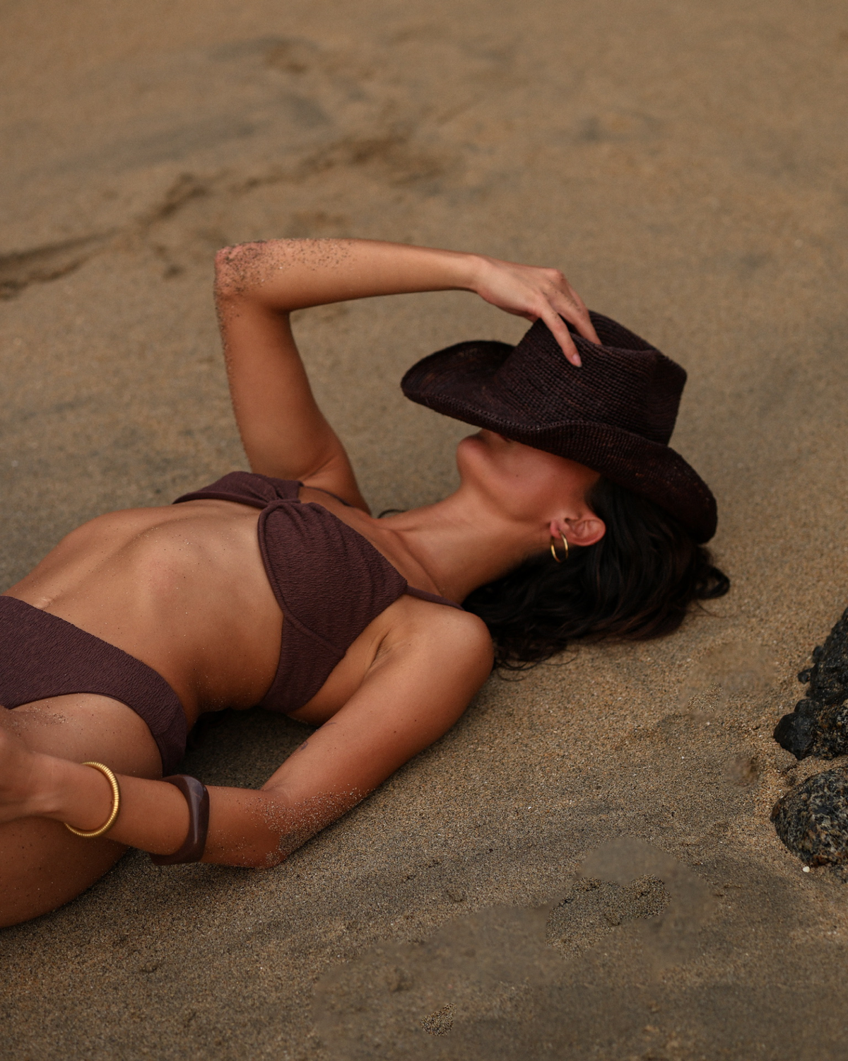 A woman lying on sand wearing a dark brown montreal bikini and a matching wide-brim hat, with her arm raised to shield her face from the sun.
