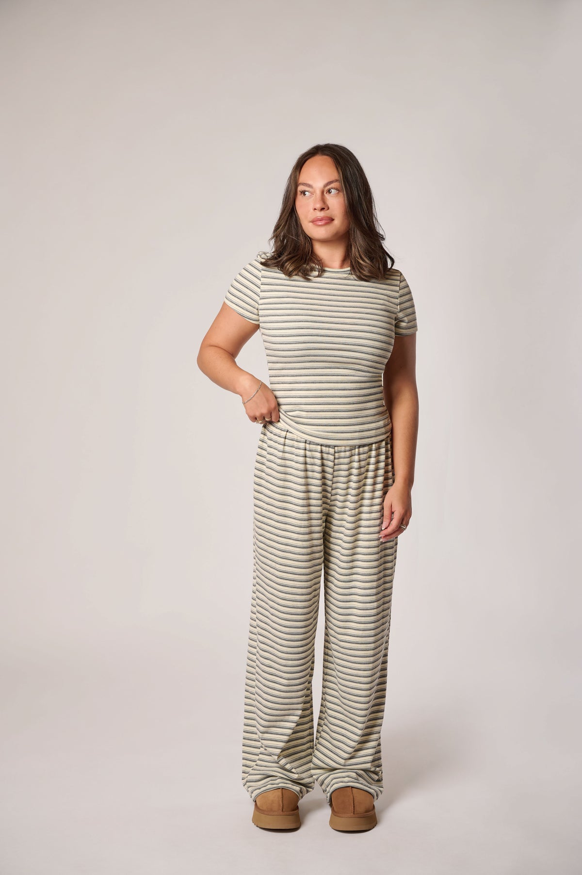 A woman modeling a matching two-piece loungewear set consisting of a short-sleeve striped t-shirt and wide-leg striped pants, paired with tan slippers, against a solid light gray studio background.
