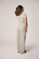 A woman is shown from the back wearing a short-sleeve with thin horizontal stripes in light and dark gray, paired with brown slippers, against a plain light beige studio background.