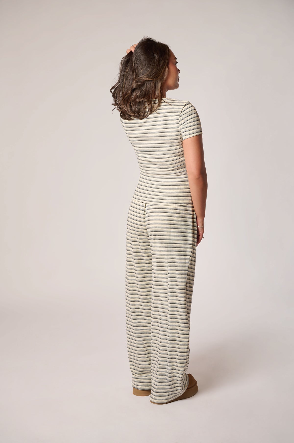 A woman is shown from the back wearing a short-sleeve with thin horizontal stripes in light and dark gray, paired with brown slippers, against a plain light beige studio background.