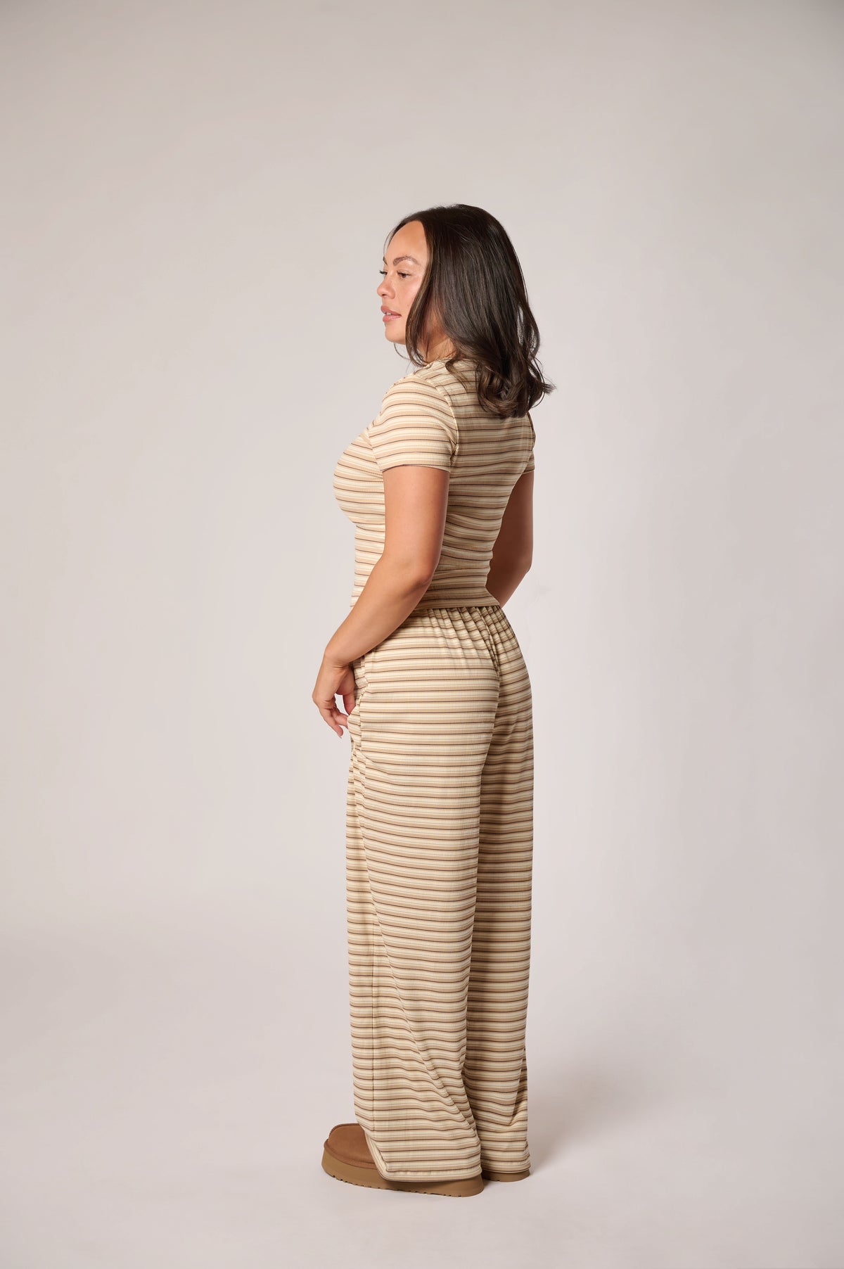 A woman stands in profile wearing a beige and brown horizontally striped short-sleeve top and matching wide-leg pants, photographed against a plain light gray studio background.