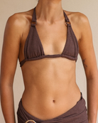 A close-up of a model wearing a dark brown crinkled bikini top with metal ring details at the shoulder straps, paired with a matching brown wrap skirt.