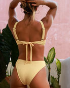 Femme portant un bikini jaune devant un mur rose avec des plantes en arrière-plan, avec un bas de maillot de couvrance brésilienne