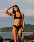 A woman modeling a black sportif two-piece bikini on a rocky beach with the ocean in the background.