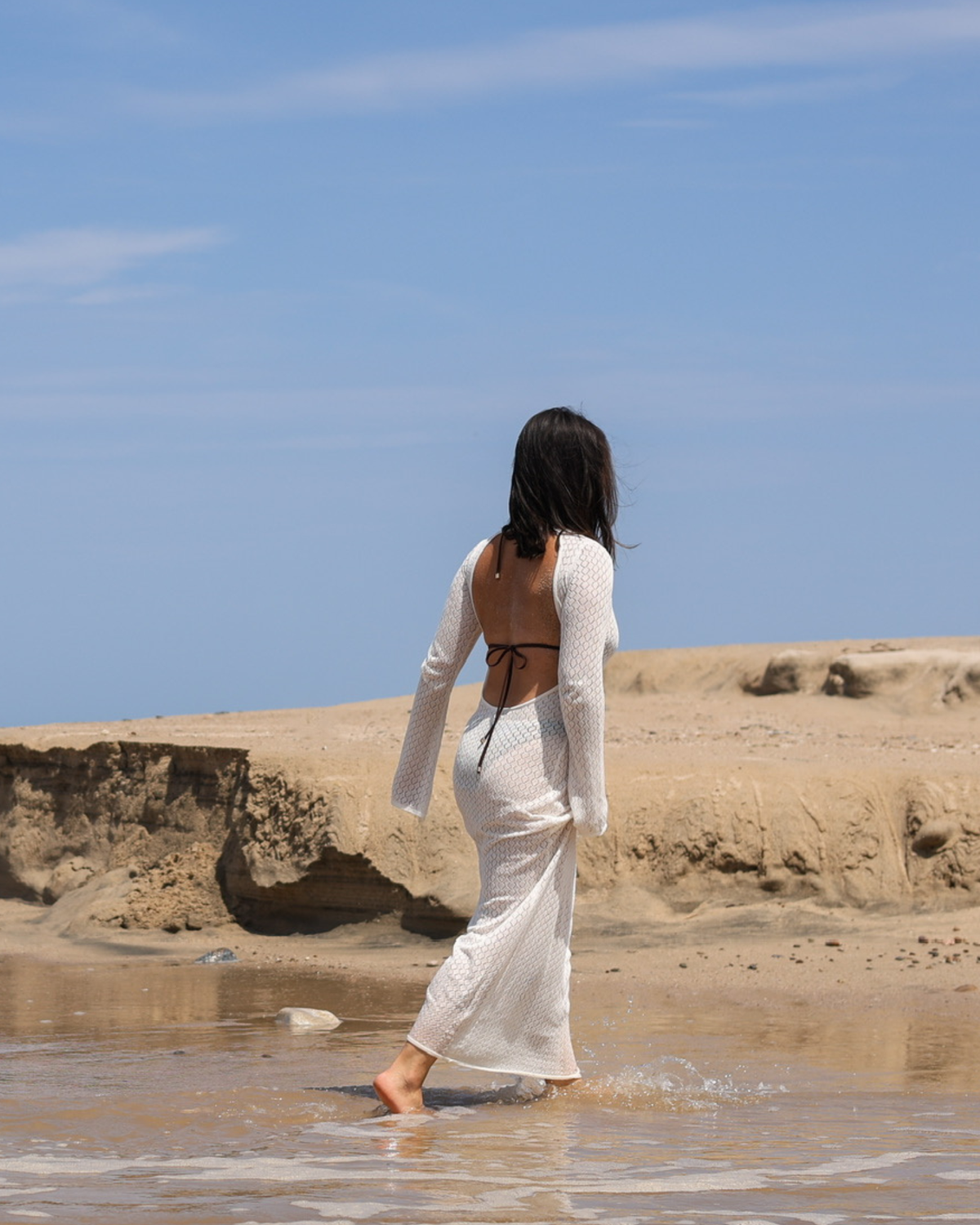 A woman stands in shallow water at a sandy beach, viewed from behind, wearing a long white open-back cover-up over a dark bikini.