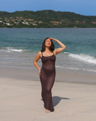 A woman wearing a dark brown knit dress stands on a sandy beach with the ocean.