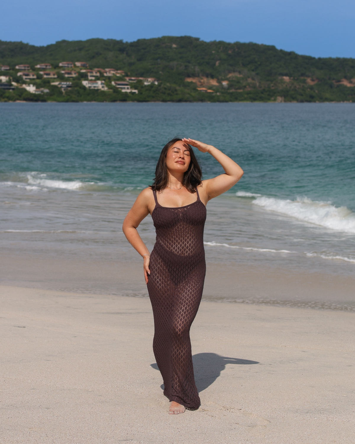 A woman wearing a dark brown knit dress stands on a sandy beach with the ocean.