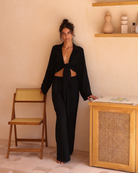 Set against warm terracotta tones, a woman stands in black pants and a black wrap top knotted at the waist, effortlessly chic.