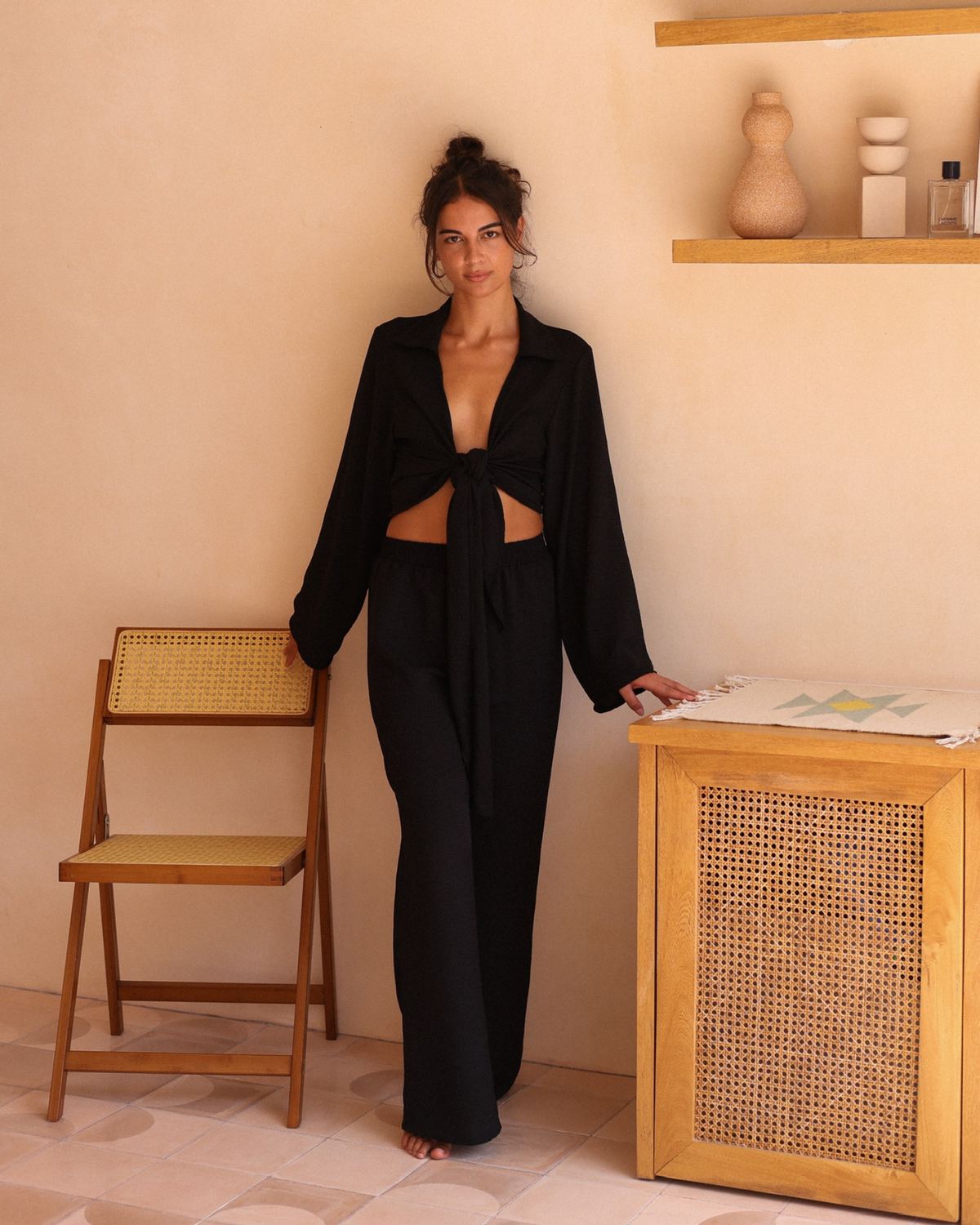 Set against warm terracotta tones, a woman stands in black pants and a black wrap top knotted at the waist, effortlessly chic.