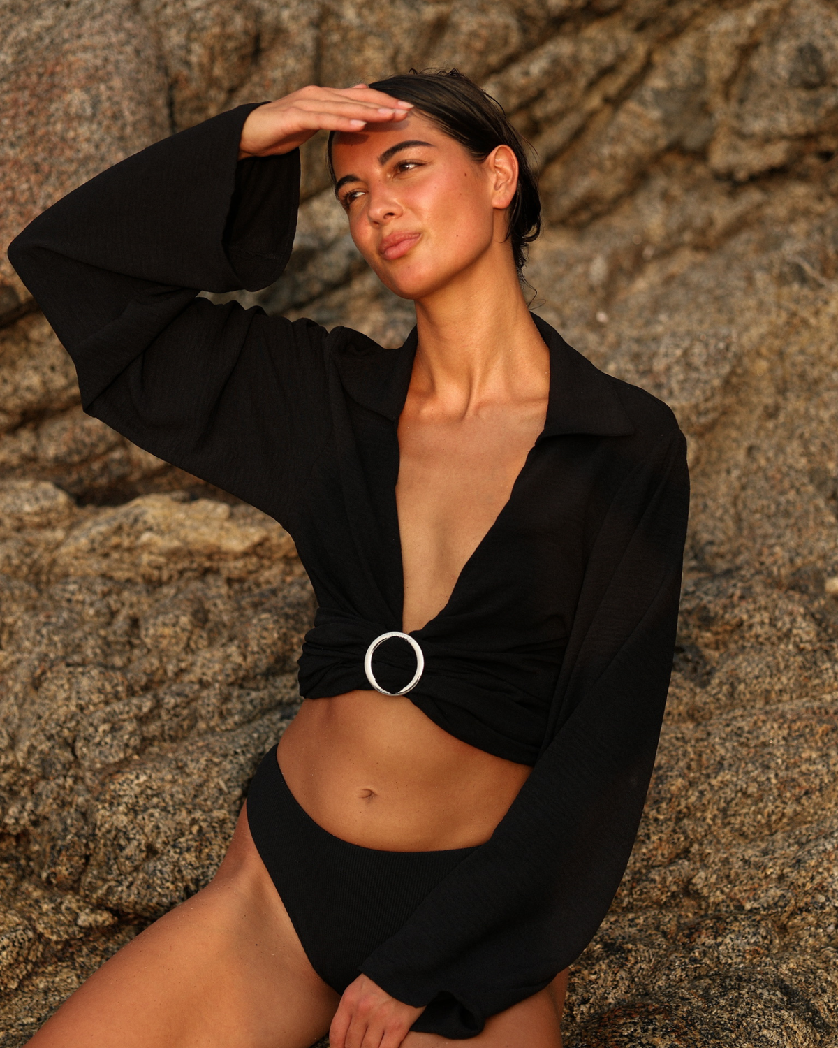 A woman posing on a rocky beach wearing a black long-sleeve crop top with a twist detail and a silver ring buckle