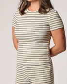 Close up of a woman wearing a short-sleeve ribbed knit top with thin horizontal stripes in beige, white, and dark gray, shown from the chest down against a plain light gray studio background.