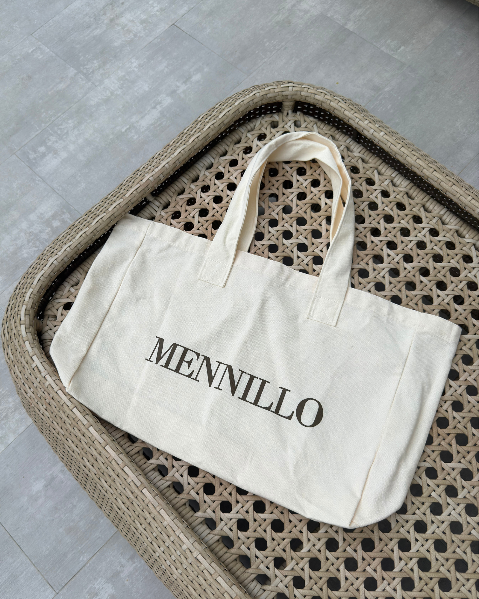 Beige tote bag with Mennillo branding, placed on a table.