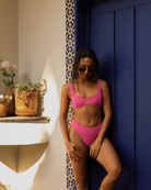 Woman in a pink bikini standing against a blue wall with potted plants and flowers in the background.